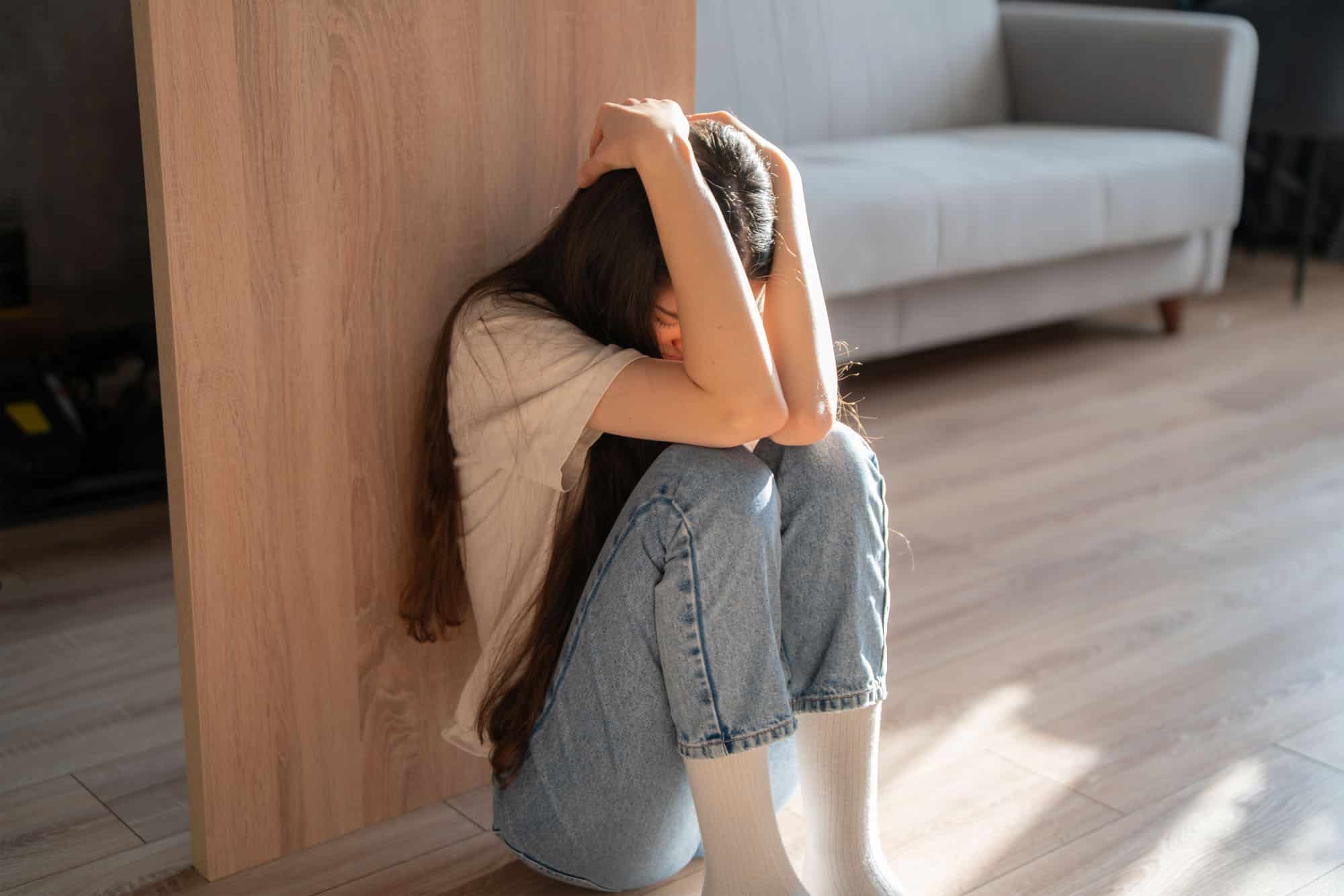 A woman sits on the floor, hugging her knees tightly while appearing lost in thought, displaying visible signs of distress in a serene environment.